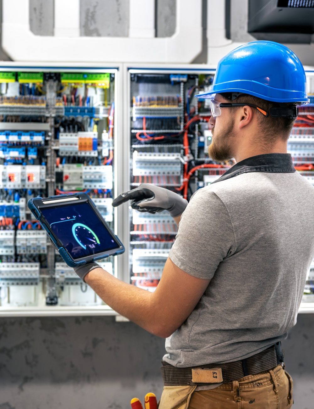 A man in protective gear reads data on a tablet in front of an electrical panel. A scene of digital system inspection in a switchboard room.